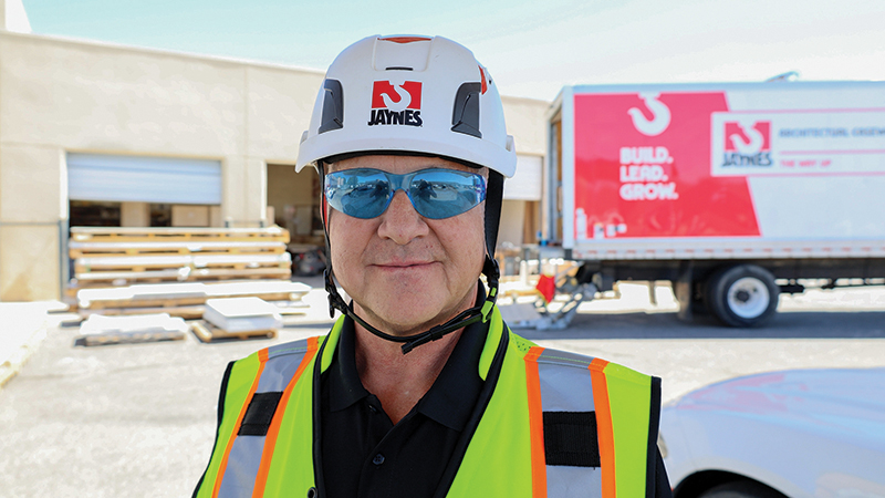 Construction worker wearing a white hard hat and reflective safety vest with Jaynes logo, standing outdoors in front of a building and a branded Jaynes truck.