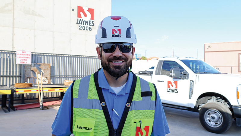 Smiling construction worker wearing a white hard hat and safety vest with the name Lorenzo, standing in front of a building, a truck, and signage bearing the Jaynes company logo.