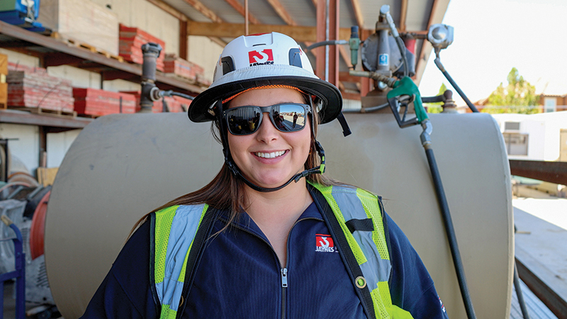 A woman wearing safety gear including a white hard hat, reflective vest, and sunglasses smiles inside an industrial or construction setting with equipment and materials in the background.