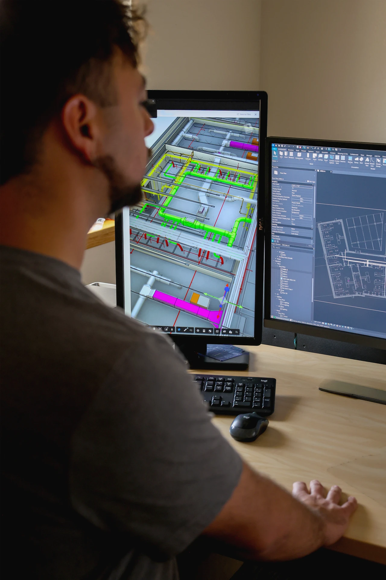 Man seated at a desk working on a computer with dual monitors displaying 3D building design and floor plan software, featuring colorful pipes and layout details.
