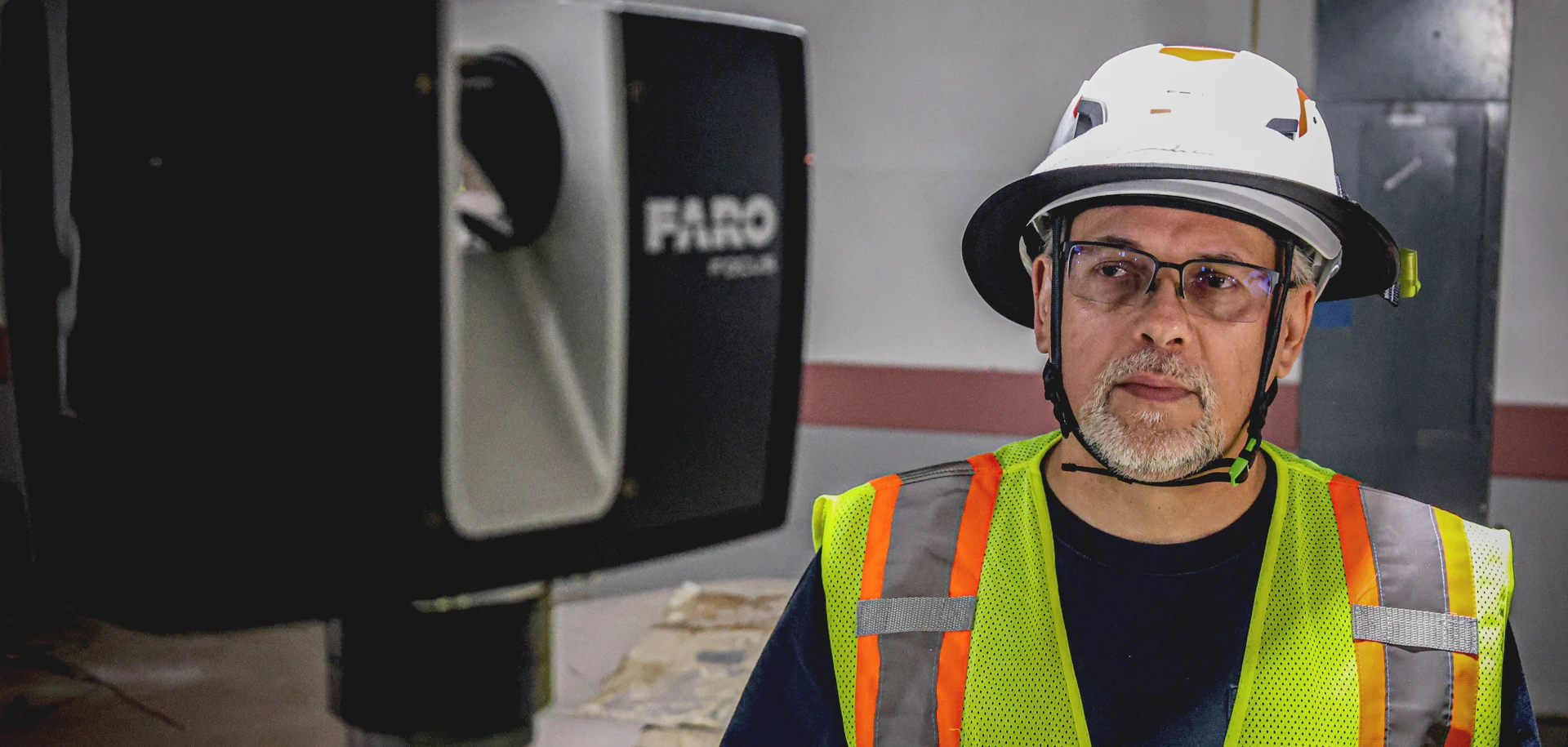 Man wearing safety helmet, glasses, and reflective yellow vest standing indoors next to a large FARO 3D scanning device.