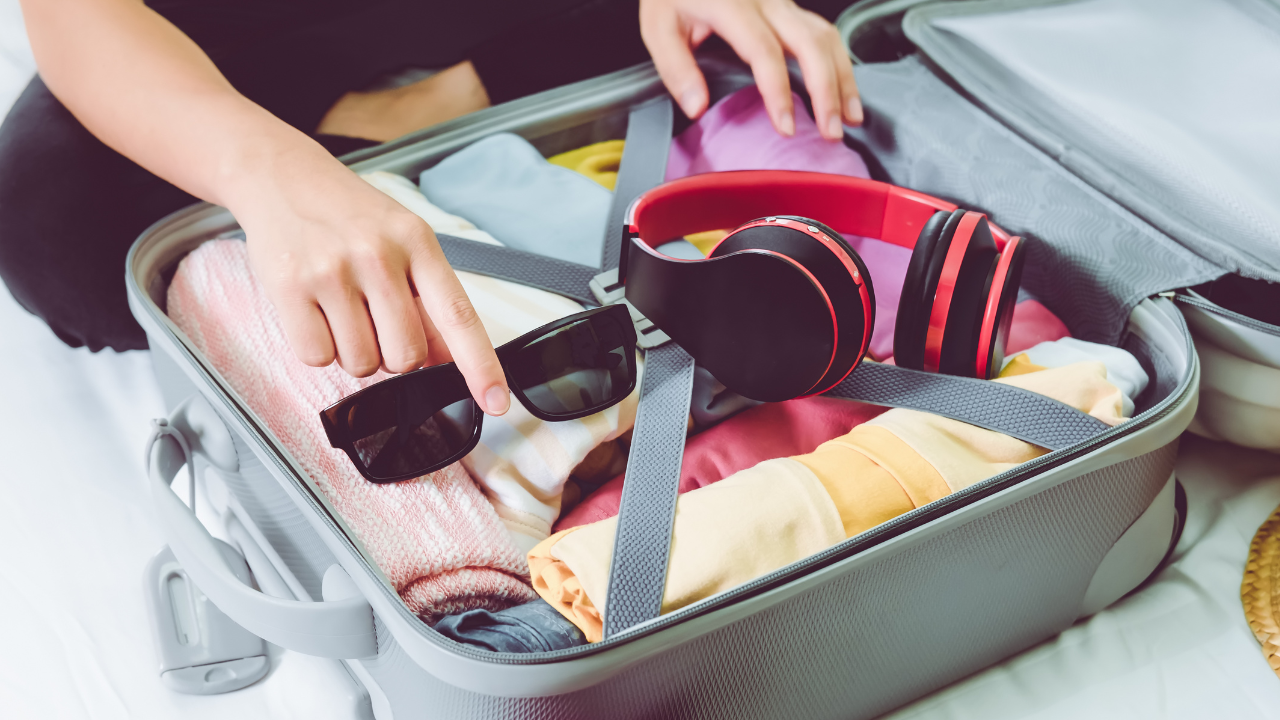 A traveler packs sunglasses, headphones, and folded clothing into an open suitcase