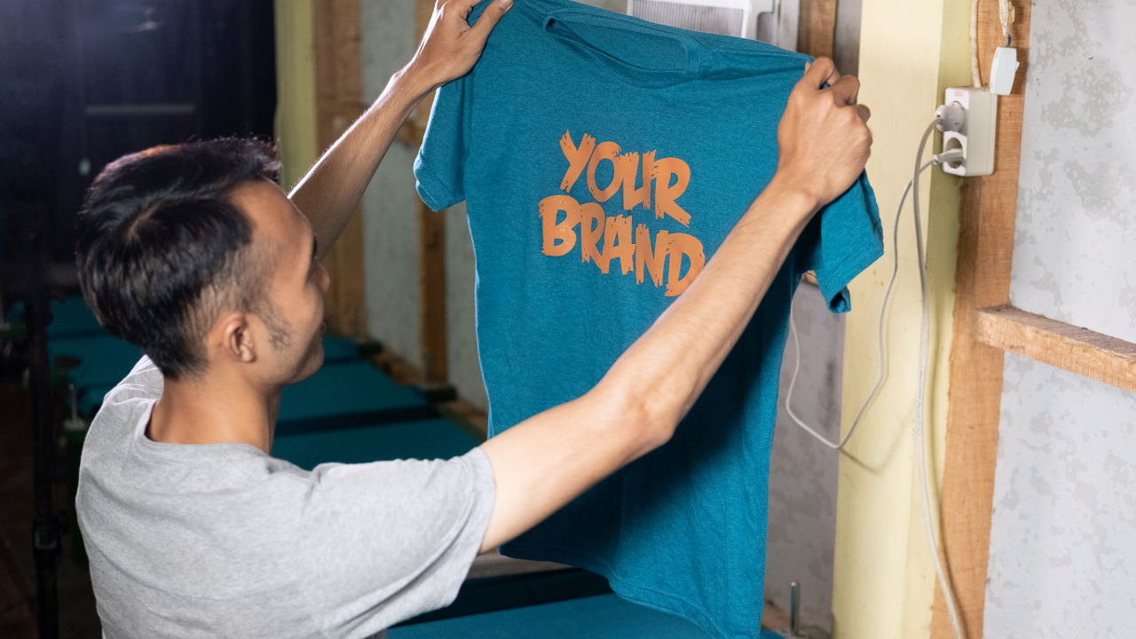 A print-shop worker holds up a blue custom T-shirt with an orange Your Brand design