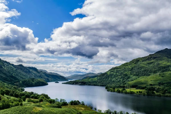 Landscape image of a crystal clear british lake surrounded by verdant hills and trees on each side and a bright blue cloudy spotted sky