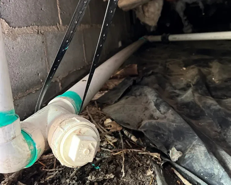 White PVC drainage pipe with green joints installed in a dirt crawl space with concrete block wall and black plastic ground cover.