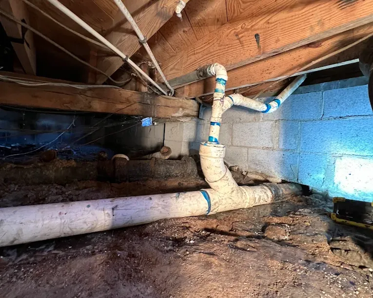 White PVC pipes with blue tape installed in a wooden-beamed crawlspace with soil floor and concrete block walls.