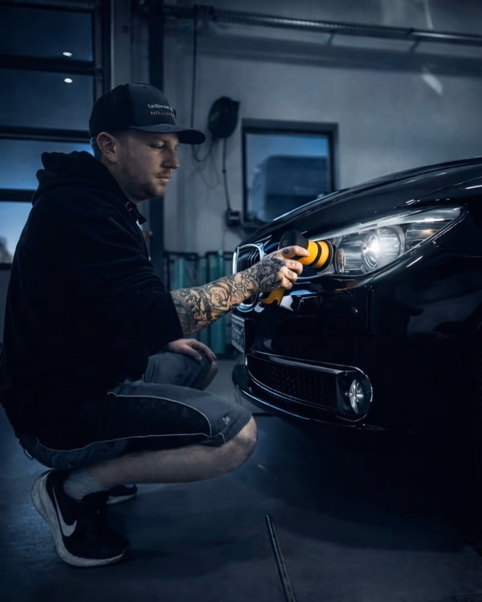 Mann mit Tätowierungen poliert Scheinwerfer eines schwarzen Autos in einer Garage.