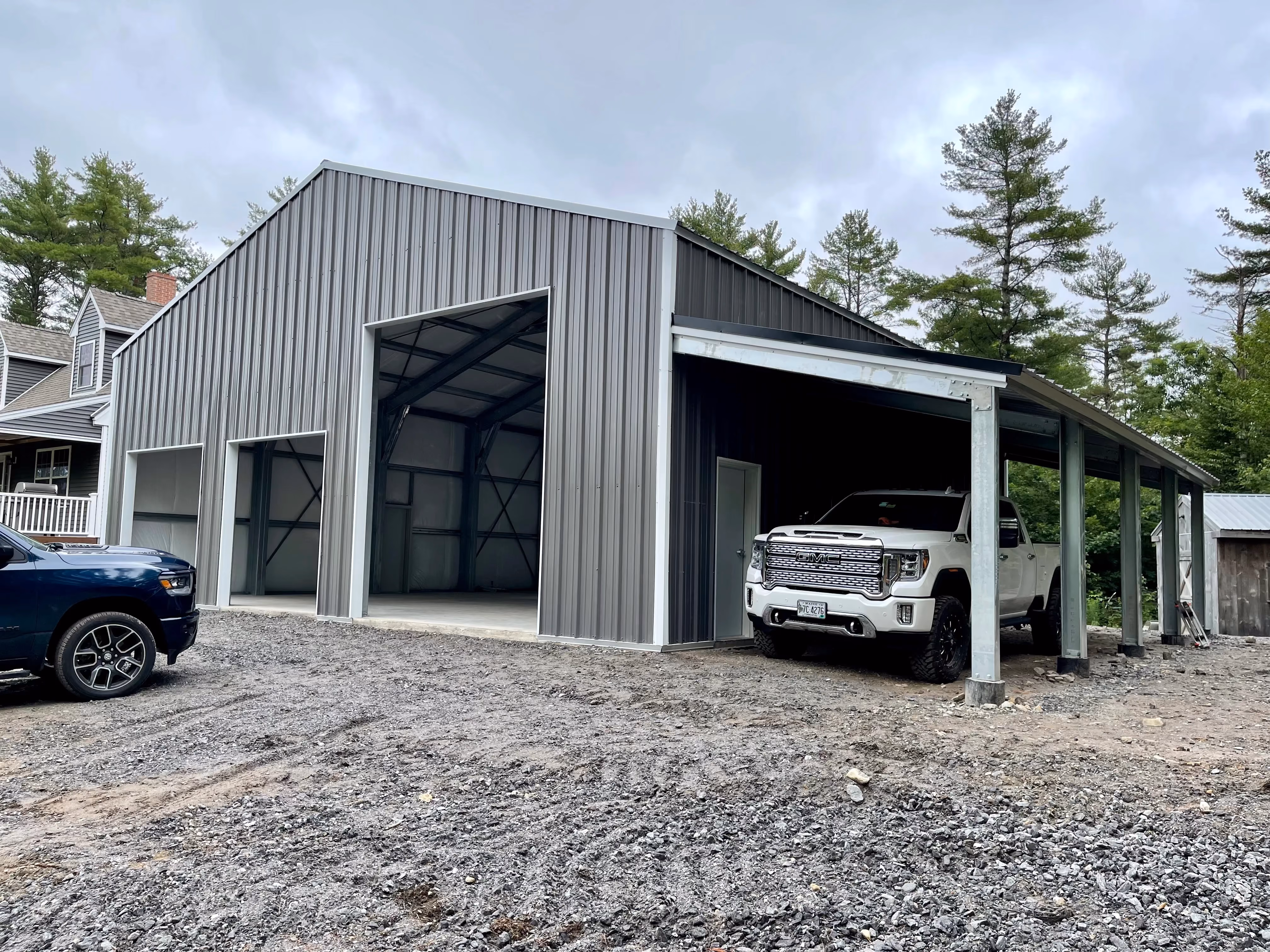 Metal three-bay steel garage with an attached carport and vehicles parked in front on a gravel driveway.