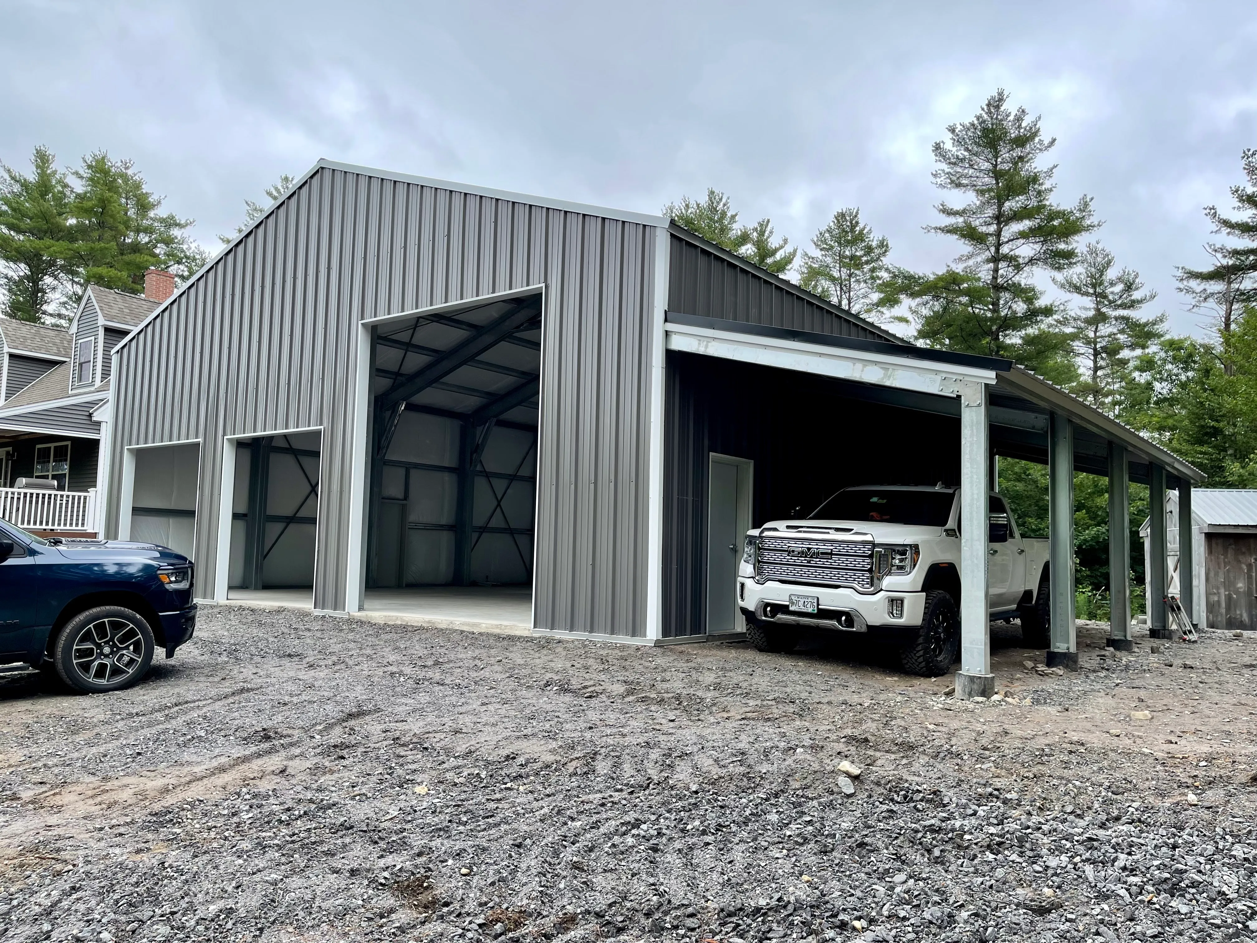 Metal three-bay steel garage with an attached carport and vehicles parked in front on a gravel driveway.