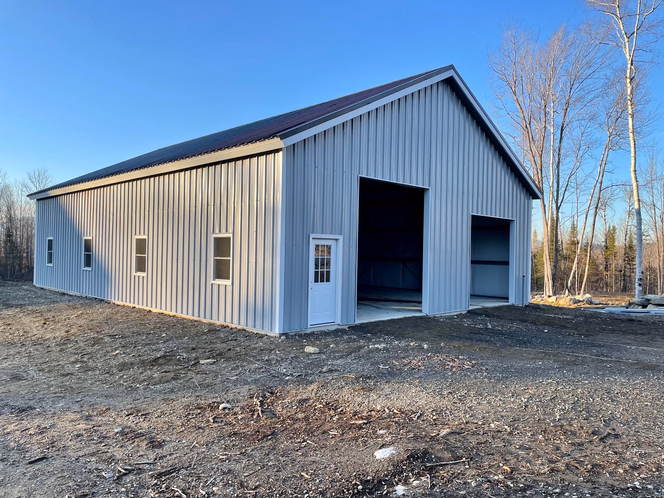 Large light gray metal building with windows and two open garage entrances on a cleared lot under a blue sky.