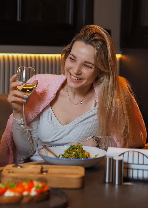 Smiling woman with long blonde hair sitting at a table holding a glass of white wine, with a bowl of pasta and appetizers in front of her.