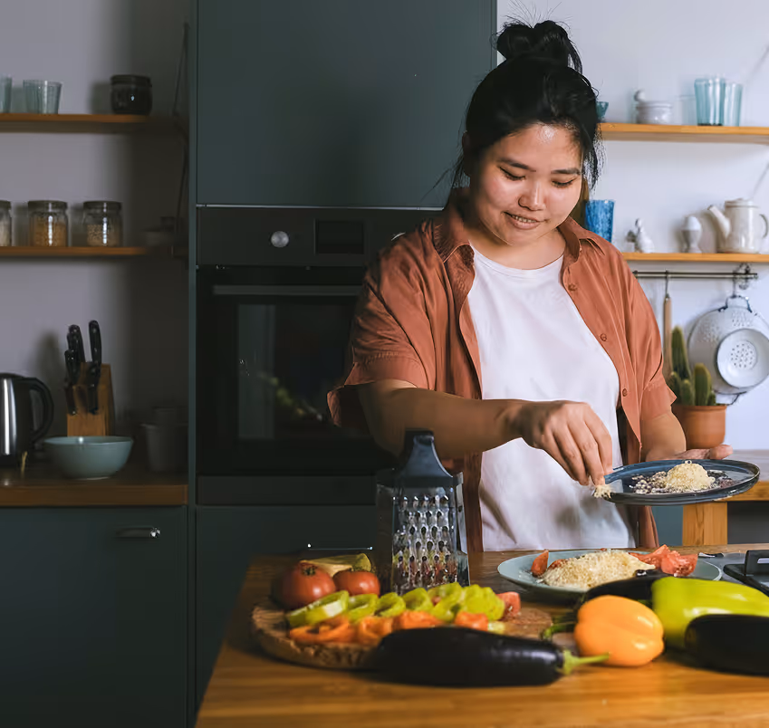 Woman sprinkling cheese on a dish in a kitchen with fresh vegetables on the counter and a box listing weekly action items for healthy habits.