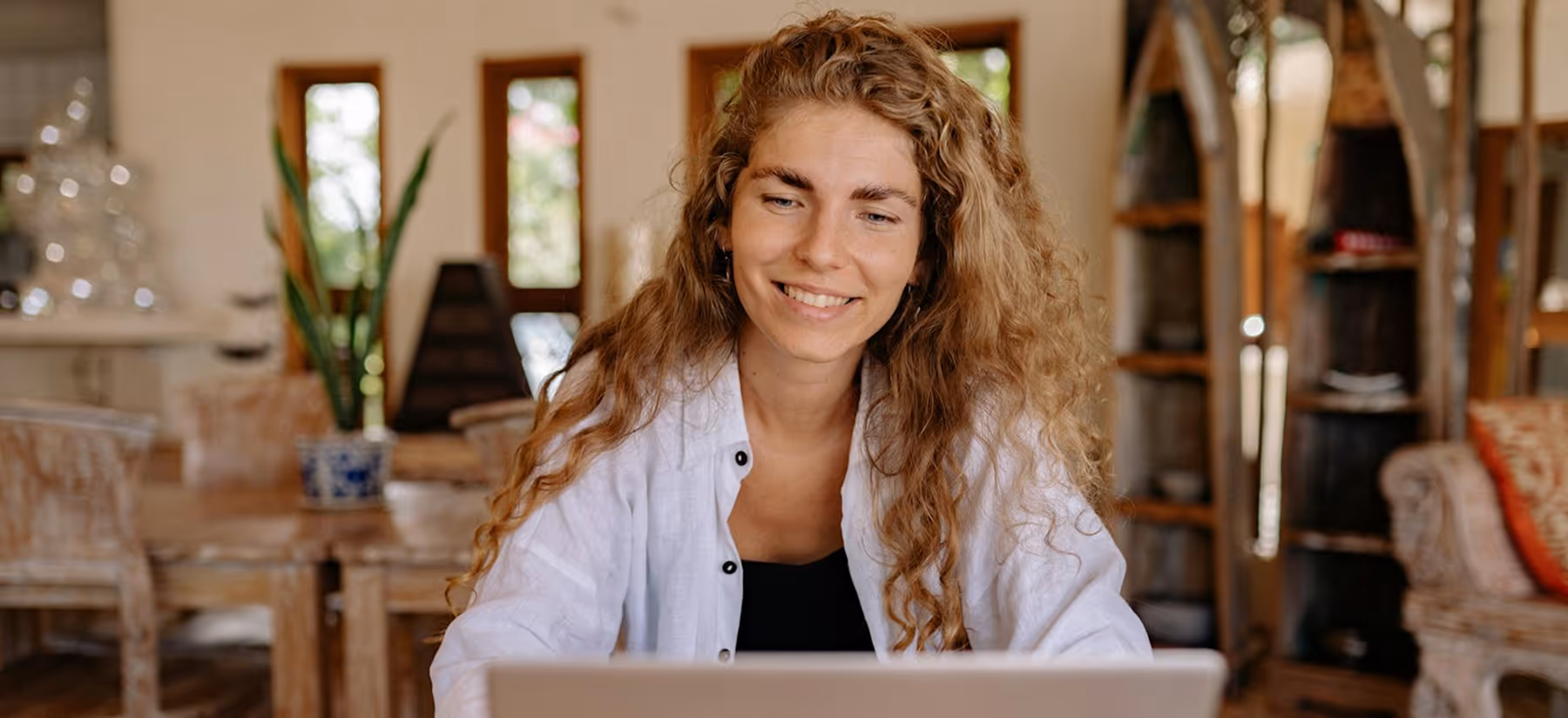 Smiling woman with curly hair using a laptop in a cozy, warmly lit room with wooden furniture.
