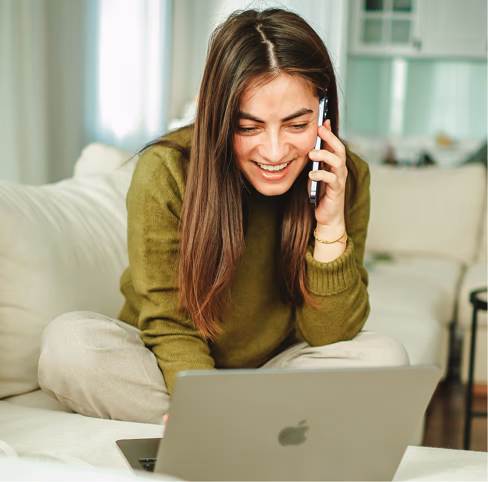 Smiling woman talking on a smartphone while looking at an open Apple laptop on a couch.