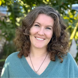 Woman with curly dark hair smiling in front of a wooden background with purple lighting on her face.