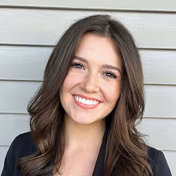 Smiling young woman with long brown hair wearing a dark blazer, standing in front of a light gray wooden wall.