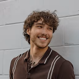 Young man with curly hair smiling and looking sideways wearing a brown shirt with beige stripes, standing against a light gray wall.