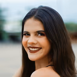 Smiling young woman with long brown hair wearing a teal top outdoors with green foliage background.