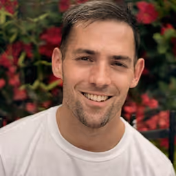 Smiling man with short brown hair wearing a white shirt, with blurred red flowers in the background.