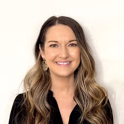 Smiling young woman with long dark hair wearing an off-shoulder black top against a beige background.