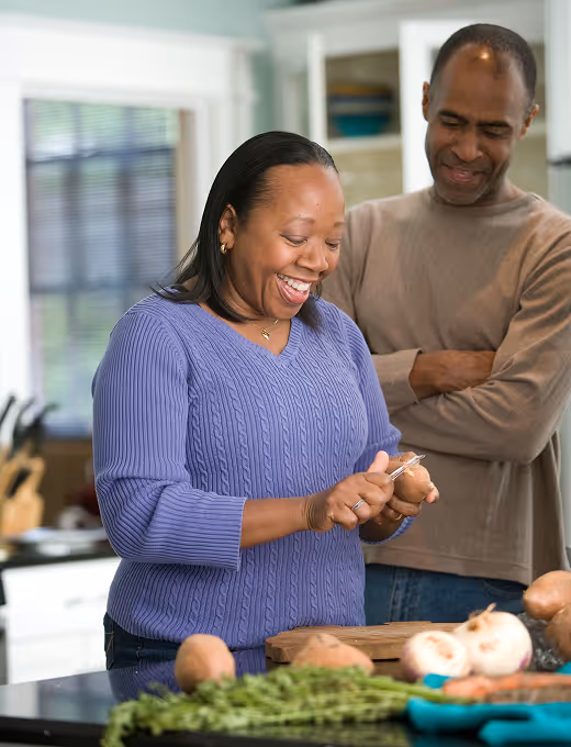 Woman peeling a potato with a knife in a kitchen while a man smiles watching her.
