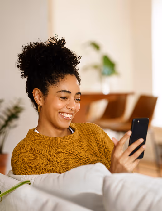 Smiling woman with curly hair in a mustard sweater looking at her smartphone while sitting on a couch.