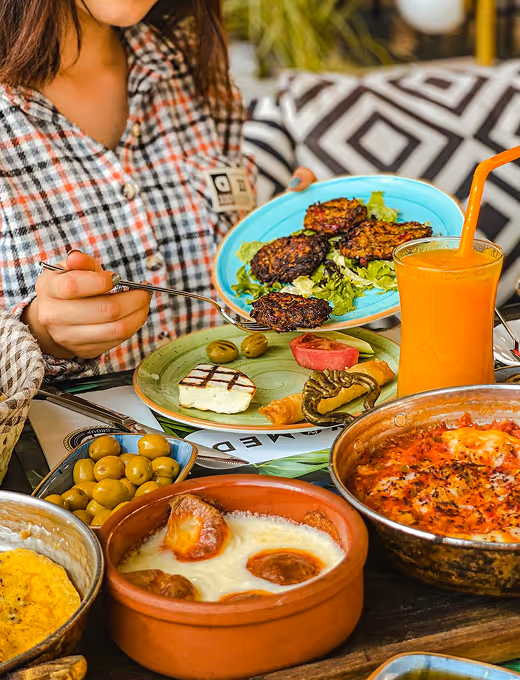 Person holding a blue plate with fried patties over a table filled with Mediterranean dishes including olives, grilled cheese, stuffed peppers, and orange juice with a straw.
