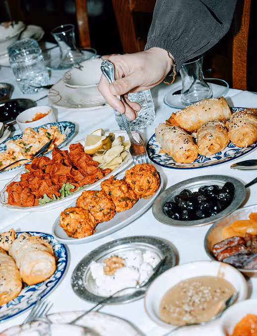 A hand holding a fork over a variety of Mediterranean dishes on a table, including kibbeh, stuffed pita breads, olives, and dips.