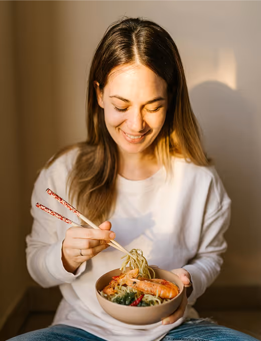 Smiling woman holding a bowl of noodles with shrimp using chopsticks.