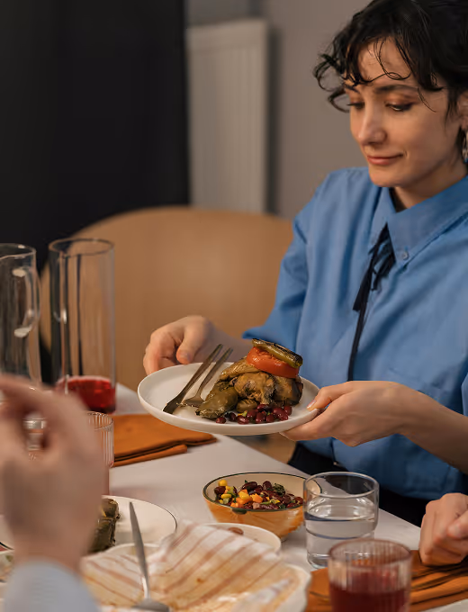 Person in blue shirt holding a plate with stuffed grape leaves and a roasted tomato, sitting at a table with drinks and food.