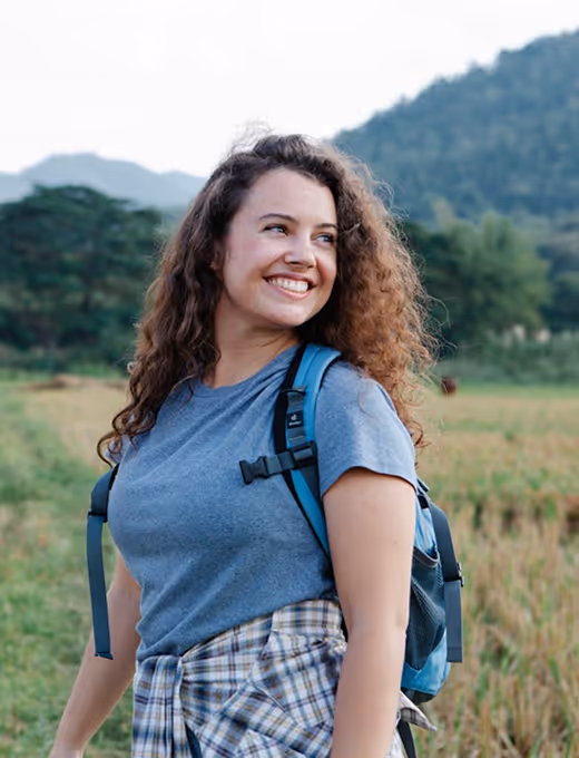 Smiling woman with curly hair wearing a blue t-shirt and a backpack standing in a field with hills in the background.