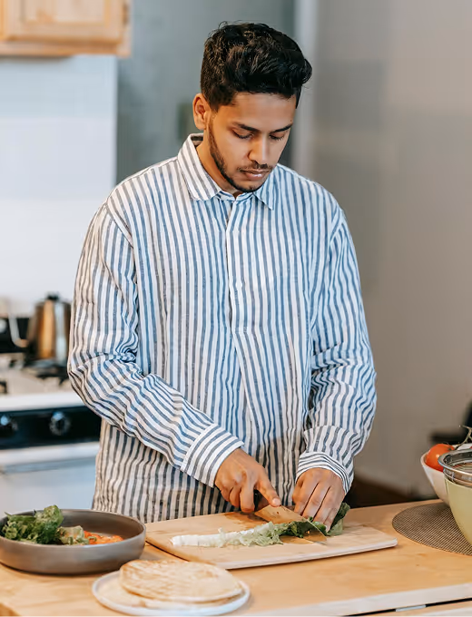 Young man in a striped shirt chopping leafy greens on a wooden cutting board in a kitchen.