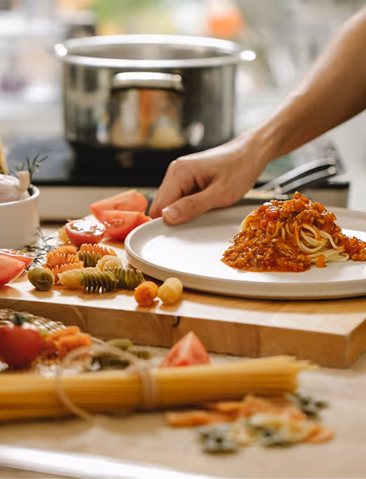 Person plating spaghetti with tomato meat sauce on a white plate in a kitchen setting with various pasta and sliced tomatoes on a wooden board.