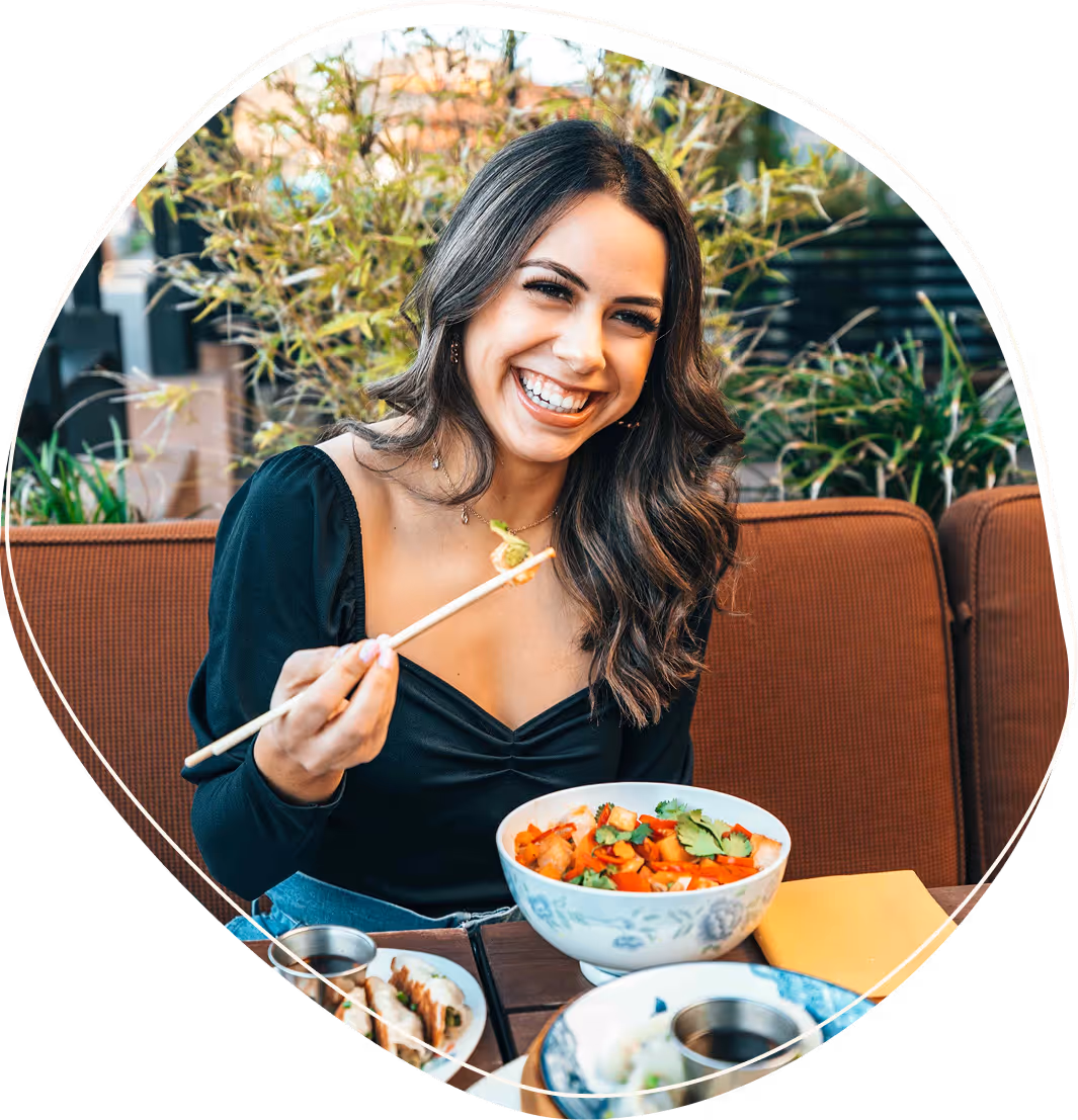 Smiling woman holding chopsticks with food, seated at table with bowls of colorful Asian cuisine.