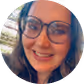 Smiling woman wearing glasses with dark hair, outdoors near a windowed structure.