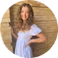 Smiling woman with long curly blonde hair wearing a white off-the-shoulder top standing in front of a wooden background.
