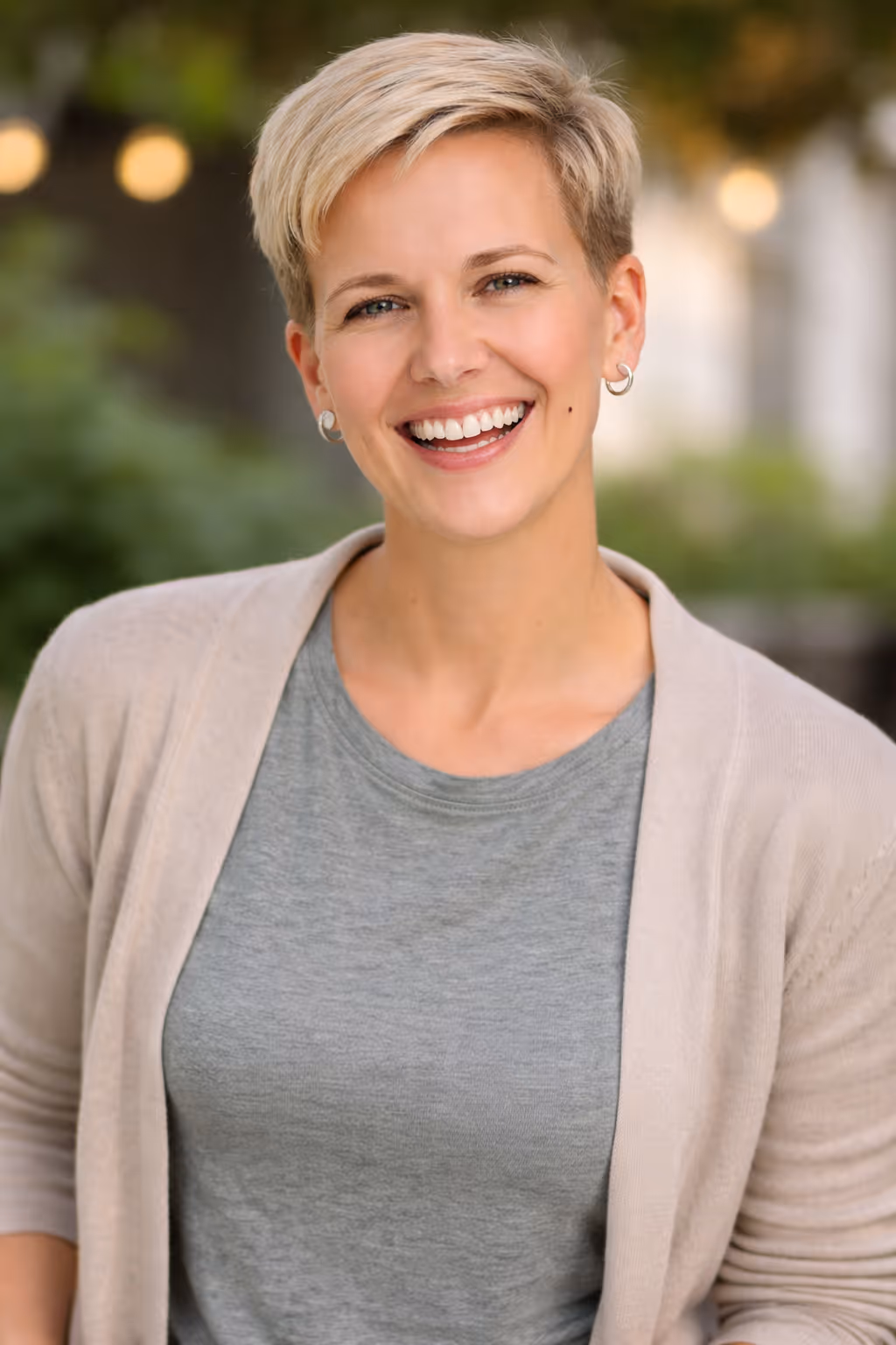 Smiling woman with short blonde hair wearing a gray shirt and beige cardigan with blurred outdoor background.