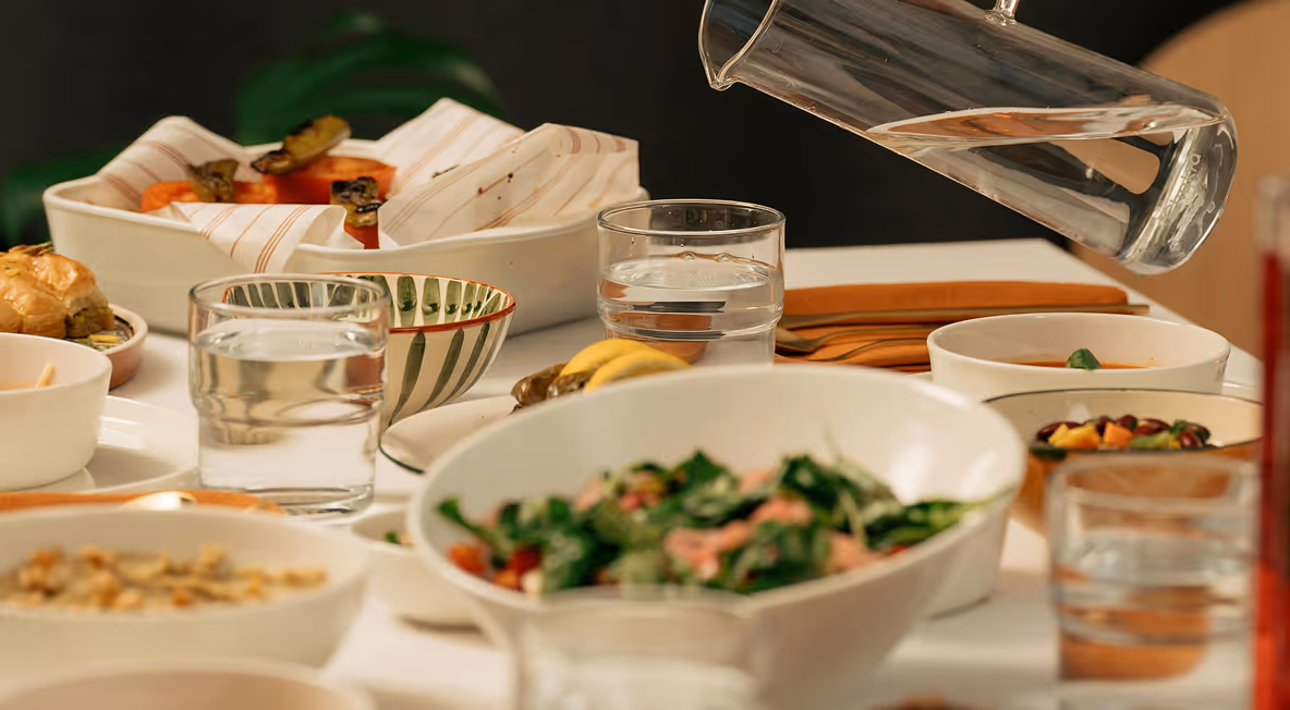 Table set with various bowls of food including salad, grilled vegetables, bread rolls, and glasses of water being poured from a pitcher.
