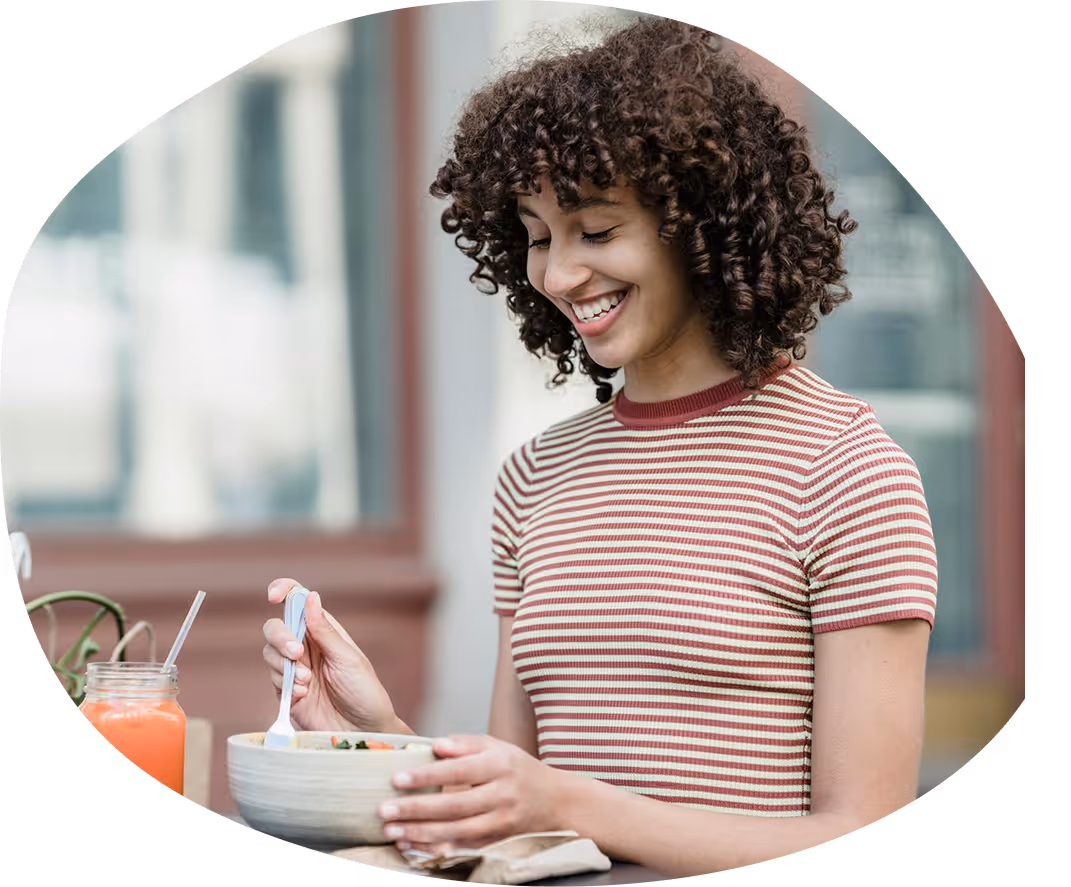 Smiling woman with curly hair wearing a striped shirt, holding a bowl and spoon while eating outdoors.