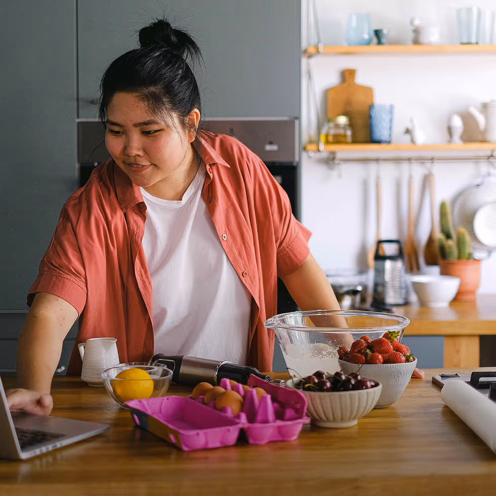 Woman in a red shirt using a laptop in a kitchen with bowls of strawberries, cherries, eggs, and a lemon on the countertop.