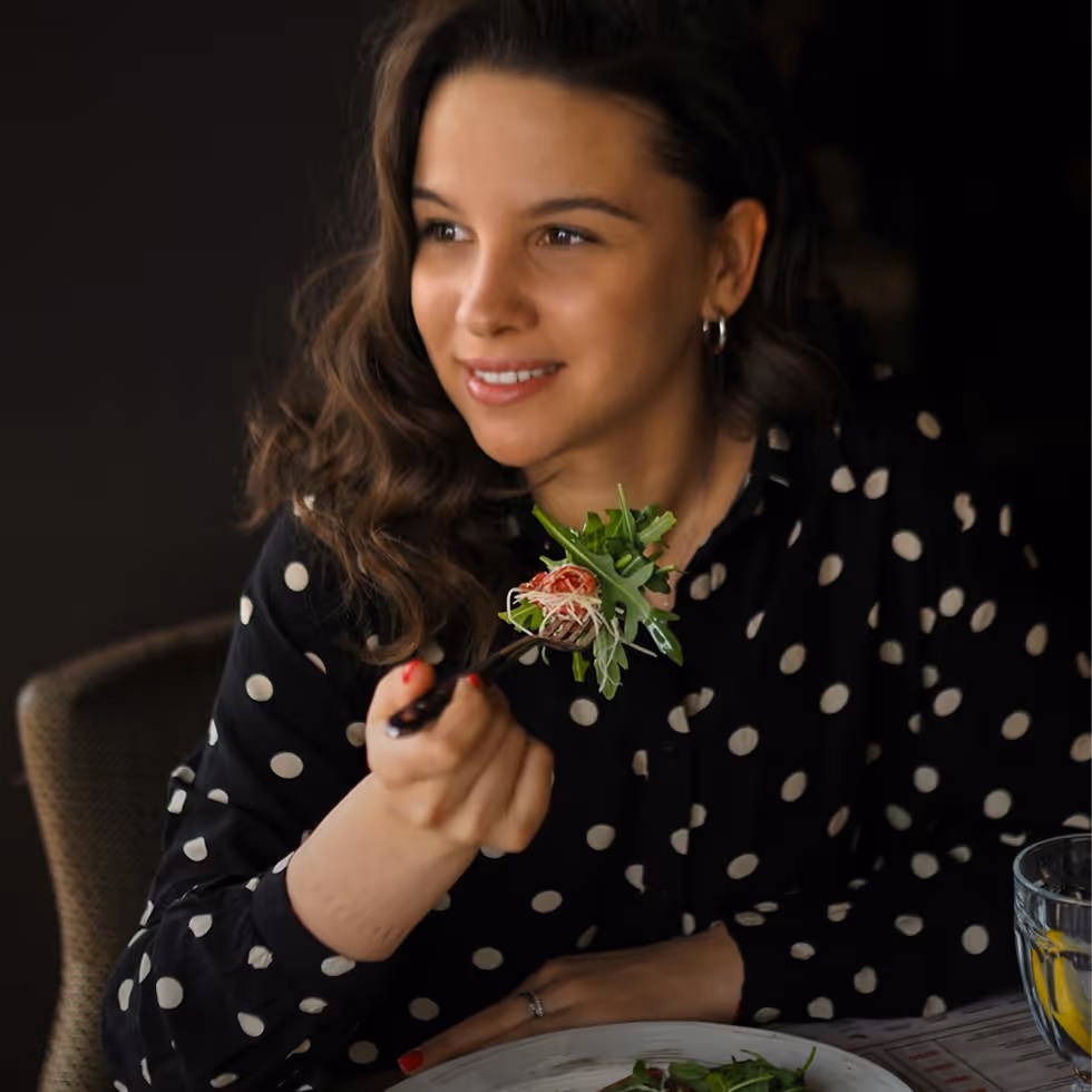 Smiling woman in black polka dot blouse holding a fork with salad greens and cherry tomato.