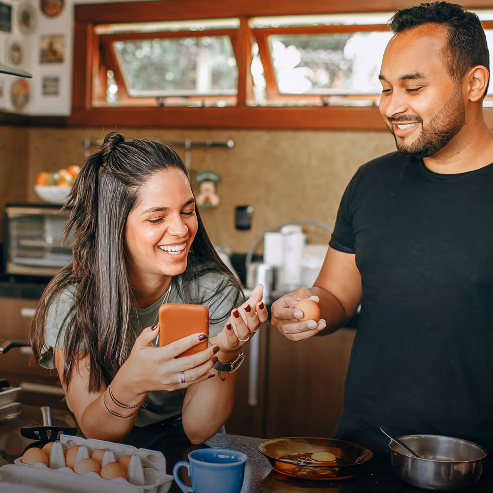 Smiling man and woman cooking in a kitchen, with the woman holding a phone and the man holding an egg over a pan.