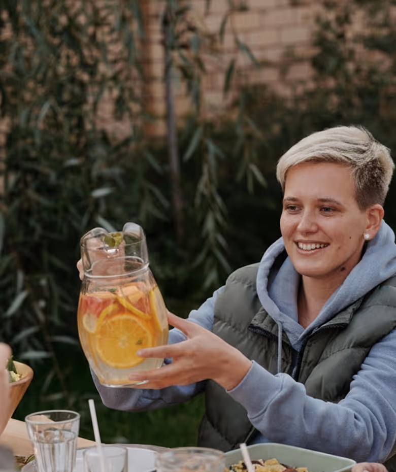 Person with short blonde hair and a gray hoodie holding a glass pitcher with citrus-infused water, smiling outdoors.