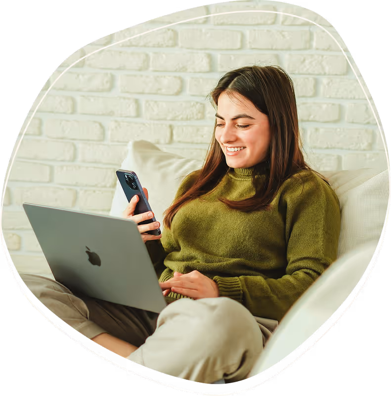 Smiling woman in a green sweater sitting on a couch using a laptop and holding a smartphone.