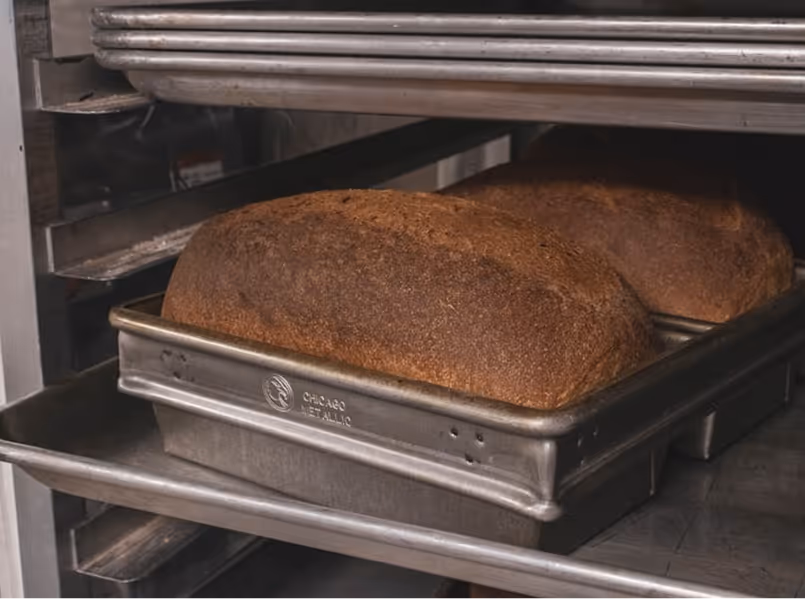 Two freshly baked brown bread loaves in metal pans inside an industrial oven.