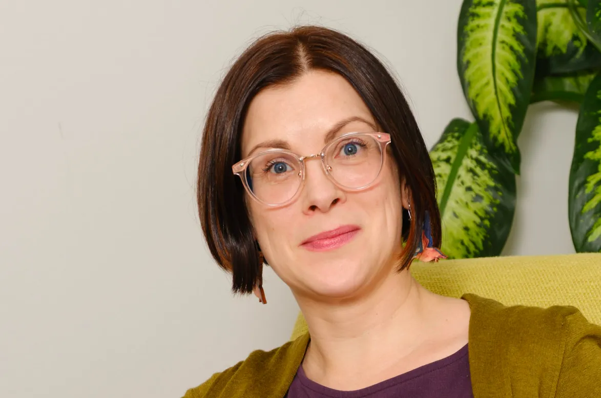 Woman with short brown hair wearing clear-framed glasses, colorful earrings, a purple top, and olive cardigan sitting in front of green leafy plant.