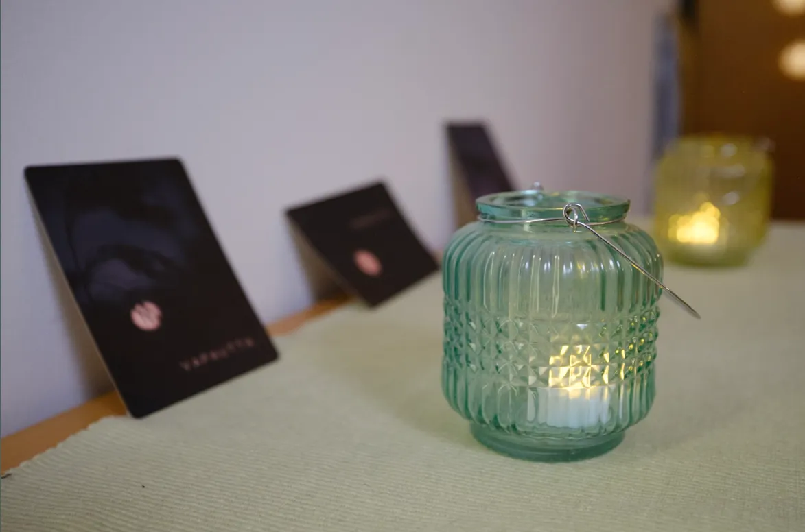 Green textured glass lantern with lit candle inside placed on a light green table runner with blurred black cards and another lantern in the background.
