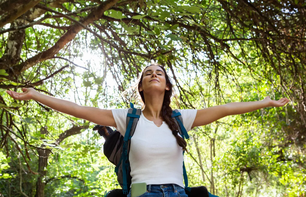 Woman with a braid wearing a backpack, standing in a sunlit forest with arms outstretched and eyes closed, enjoying nature.
