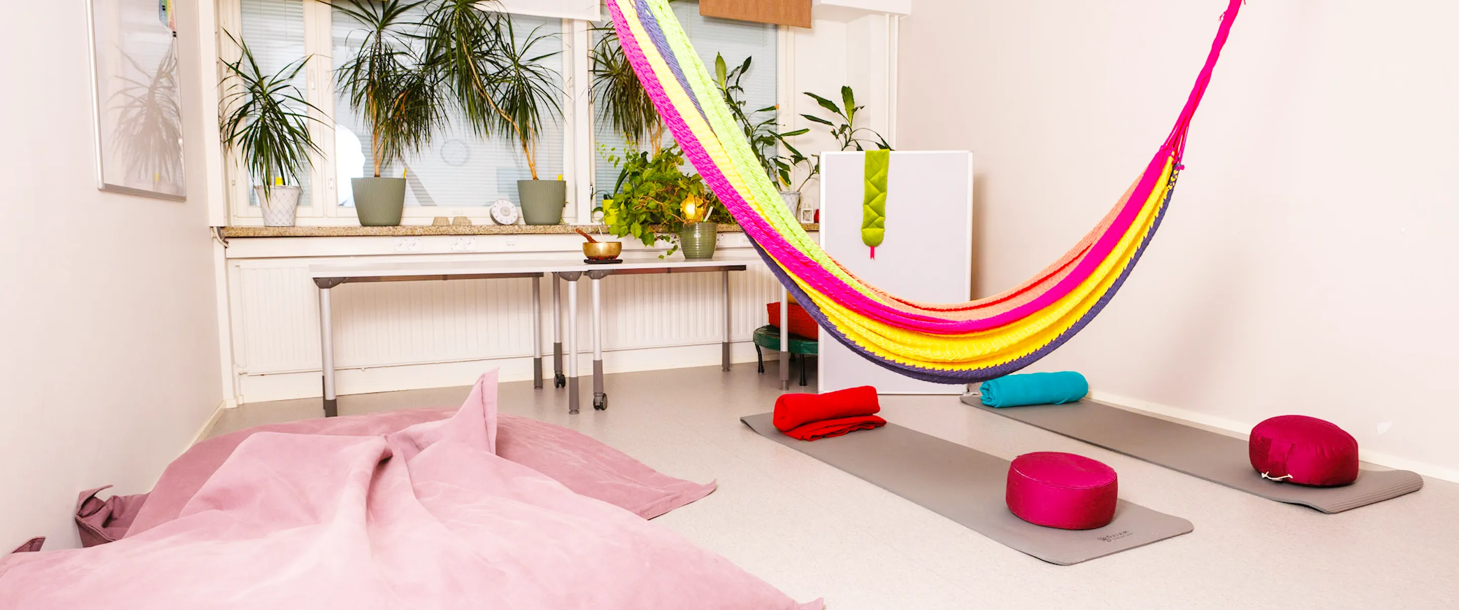 Bright room with large window, colorful hammock, yoga mats with rolled towels and meditation cushions, and several potted plants on the windowsill and floor.