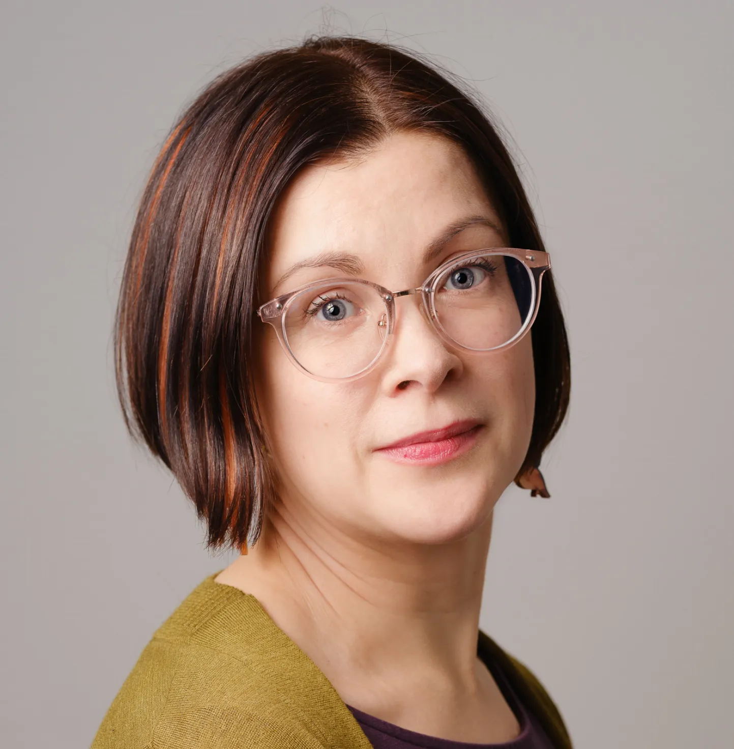 Woman with short brown hair, wearing clear round glasses and a green sweater, looking at the camera against a gray background.
